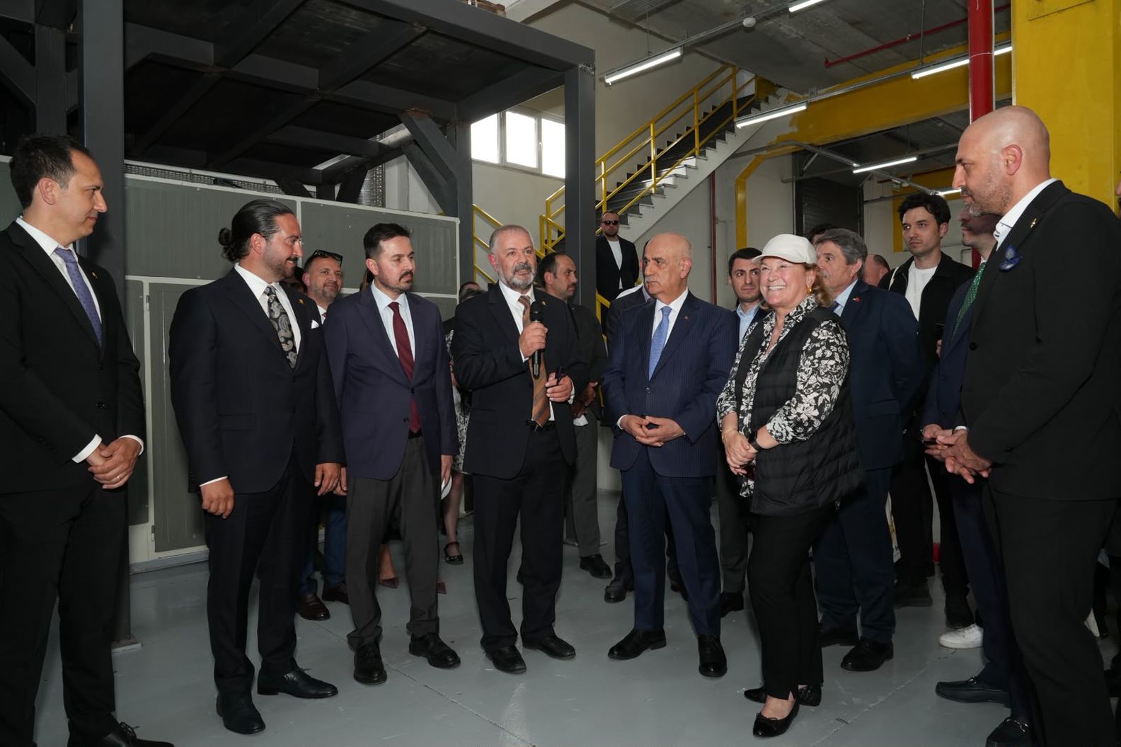 Tesisin içinde mikrofonla konuşan bir kişiyi dinleyen kalabalık bir grup.